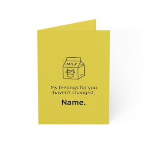 May include: A folded yellow greeting card with a black line drawing of a milk carton with a cow on it. The text on the card reads "My feelings for you haven't changed, Name."
