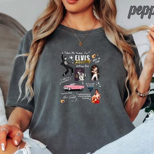 May include: A dark gray t-shirt with a graphic print of Elvis Presley. The graphic includes images of Elvis, a pink Cadillac, and a guitar. The text on the graphic includes "Love Me Tender", "Elvis Presley", "Jailhouse Rock", and "Don't Be Cruel".