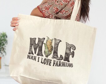 May include: Cream-colored tote bag with the text "MILF MAN I LOVE FARMING" in a distressed font. A graphic of an ear of corn is in the center of the word "MILF". The bag has a simple design with long straps for carrying over the shoulder.