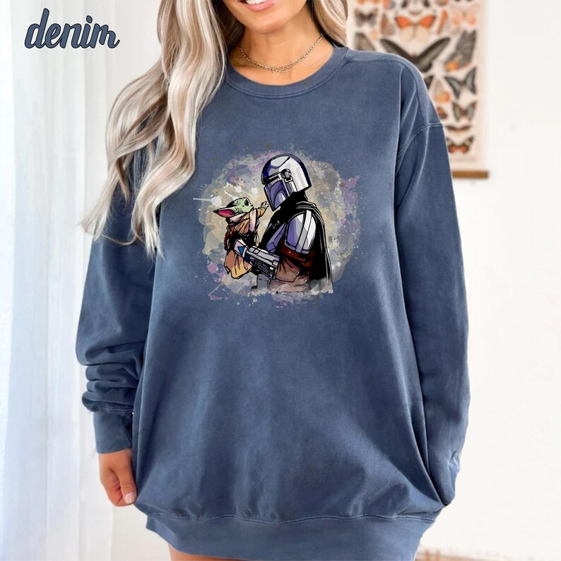 May include: A blue long-sleeved sweatshirt with a watercolor-style image of a Mandalorian character holding a small green alien creature.
