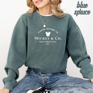 May include: A teal blue sweatshirt with a white graphic that says "Please Return to Mickey & Co. Main Street, USA 1928" with a Mickey Mouse head silhouette.