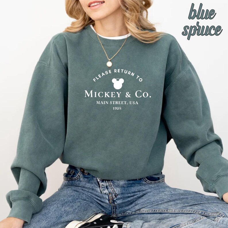 May include: A teal blue sweatshirt with a white graphic that says "Please Return to Mickey & Co. Main Street, USA 1928" with a Mickey Mouse head silhouette.