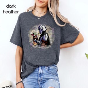 K&ouml;nnte beinhalten: Ein dunkel-heidesgraues T-Shirt mit einer Aquarell-Illustration eines Mandalorianer-Charakters, der ein kleines gr&uuml;nes au&szlig;erirdisches Wesen h&auml;lt. Die Illustration ist von einem hellblauen und violetten Aquarell-Hintergrund umgeben.