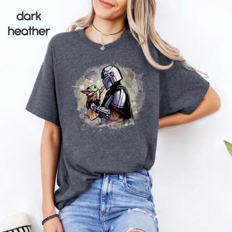 K&ouml;nnte beinhalten: Ein dunkel-heidesgraues T-Shirt mit einer Aquarell-Illustration eines Mandalorianer-Charakters, der ein kleines gr&uuml;nes au&szlig;erirdisches Wesen h&auml;lt. Die Illustration ist von einem hellblauen und violetten Aquarell-Hintergrund umgeben.