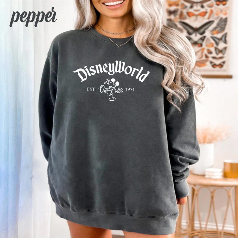 Puede incluir: Una sudadera gris oscura con un estampado gr&aacute;fico blanco de Mickey Mouse y el texto "Disneyworld Est. 1971".