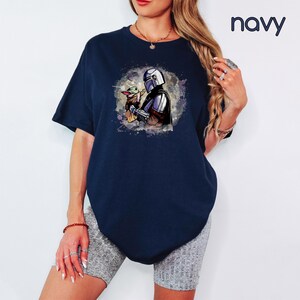 K&ouml;nnte beinhalten: Ein marineblaues T-Shirt mit einer Aquarell-Illustration eines Mandalorianer-Charakters, der ein kleines, gr&uuml;nes Wesen h&auml;lt. Die Illustration ist von einem wei&szlig;en, gesprenkelten Hintergrund umgeben.