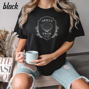 May include: Black t-shirt with a white graphic of a seashell and the text "Ariel's Swim Club Est. 1989".