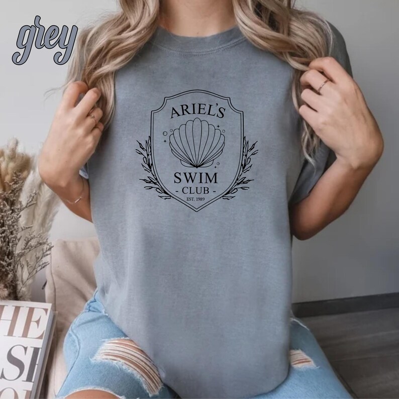 May include: A gray t-shirt with a black graphic design featuring a seashell and the text "Ariel's Swim Club - Est. 1989".