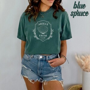 May include: A green t-shirt with a white graphic of a seashell and the text "Ariel's Swim Club Est. 1989". The shirt is worn by a person with blue denim shorts.