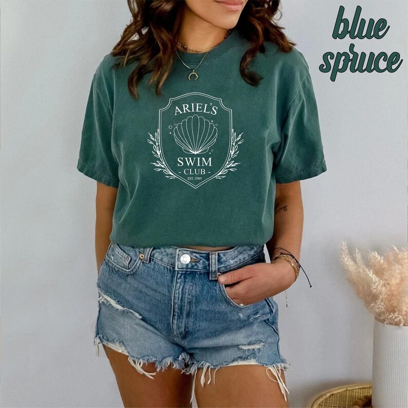 May include: A green t-shirt with a white graphic of a seashell and the text "Ariel's Swim Club Est. 1989". The shirt is worn by a person with blue denim shorts.