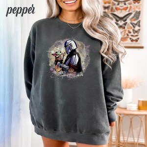 May include: A dark gray sweatshirt with a watercolor-style image of a Mandalorian character holding a small green creature. The image is centered on the sweatshirt.