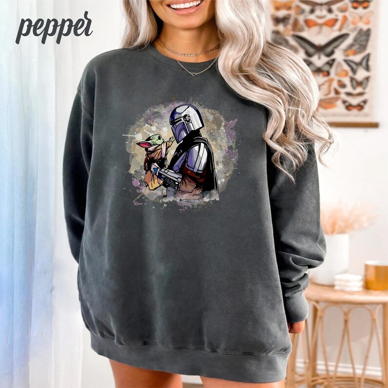 May include: A dark gray sweatshirt with a watercolor-style image of a Mandalorian character holding a small green creature. The image is centered on the sweatshirt.