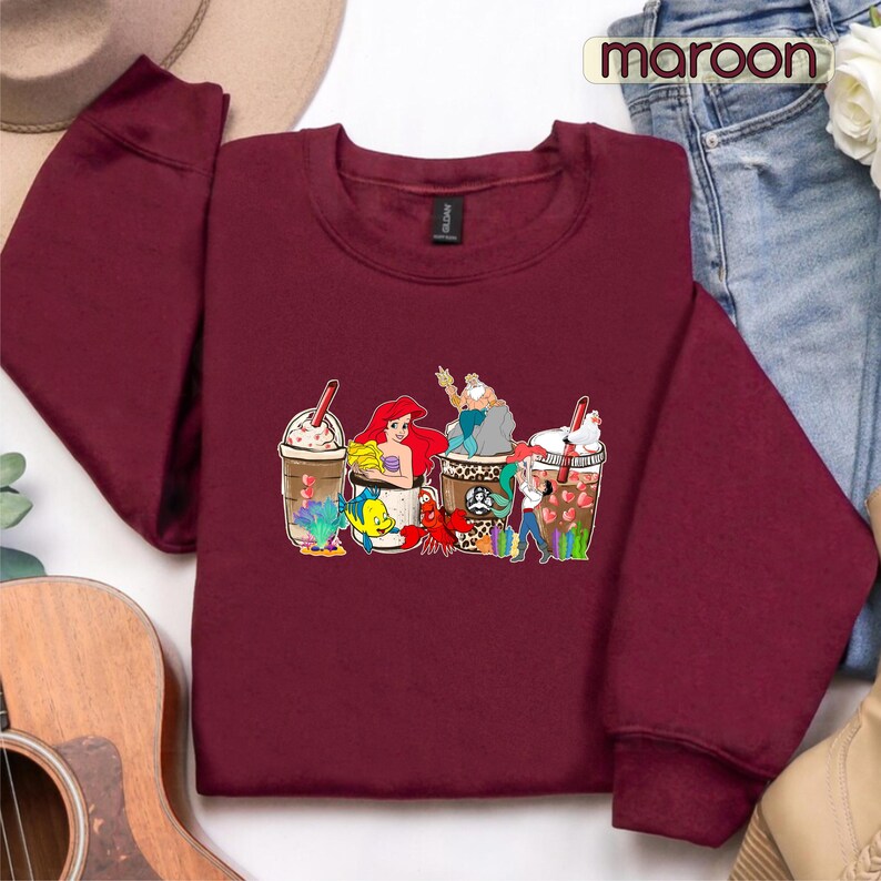 May include: A maroon sweatshirt with a graphic of four coffee cups featuring characters from the Little Mermaid. The cups are decorated with sea-themed designs, including a mermaid, a crab, a starfish, and a seashell.