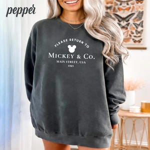 May include: A charcoal grey sweatshirt with a white graphic print that says "Please return to Mickey & Co. Main Street, USA 1928" with a Mickey Mouse head icon.