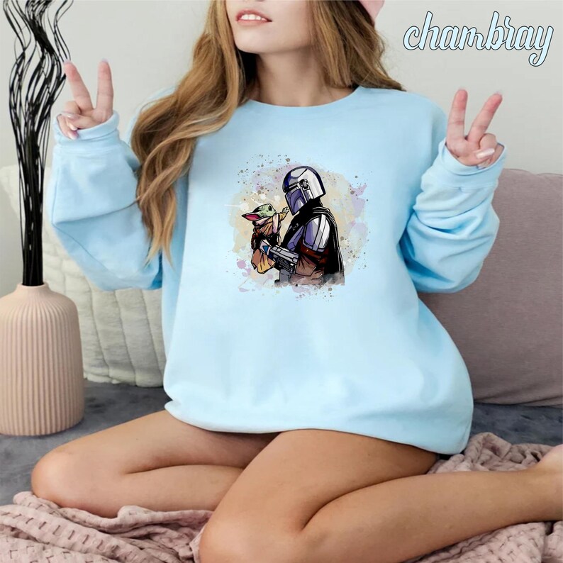 May include: A light blue sweatshirt with a watercolor-style image of a Mandalorian character holding a small, green alien creature. The image is surrounded by a light blue and pink watercolor background.