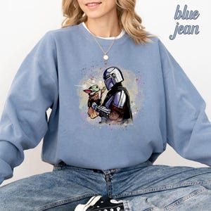 May include: A blue sweatshirt with a watercolor design of a Mandalorian character holding a small, green alien. The design is centered on the sweatshirt.