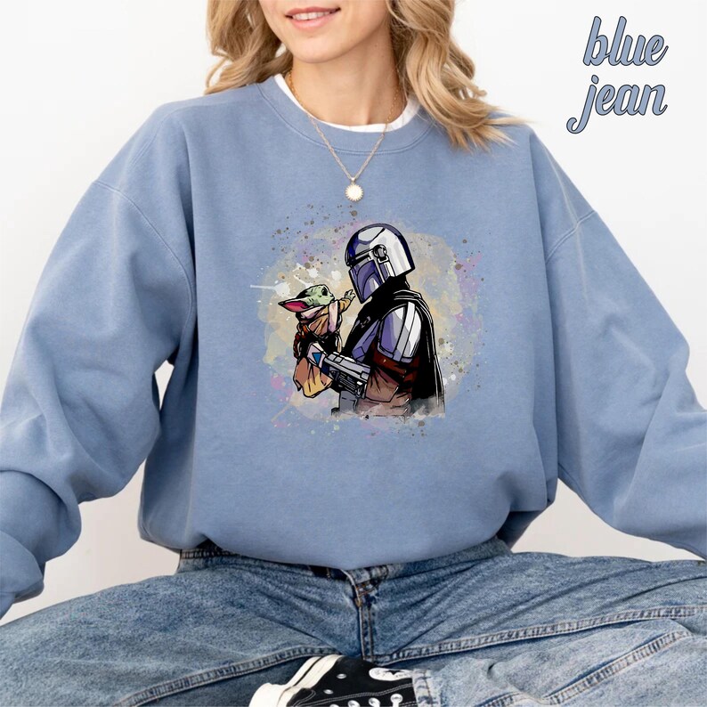 May include: A blue sweatshirt with a watercolor design of a Mandalorian character holding a small, green alien. The design is centered on the sweatshirt.