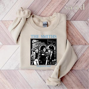 May include: A beige sweatshirt with a black and white image of the band The Smiths. The image includes the band members standing in front of a building with the text "The Queen Is Dead" below the image.