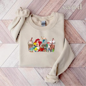 May include: A tan sweatshirt with a graphic print of four coffee cups featuring characters from the Disney movie The Little Mermaid. The cups are topped with whipped cream and chocolate drizzle. The characters include Ariel, Flounder, Sebastian, and Ursula. The text "sand" is printed in the top right corner of the image.