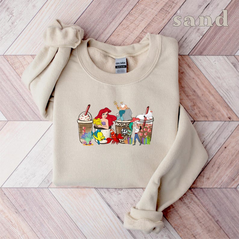 May include: A tan sweatshirt with a graphic print of four coffee cups featuring characters from the Disney movie The Little Mermaid. The cups are topped with whipped cream and chocolate drizzle. The characters include Ariel, Flounder, Sebastian, and Ursula. The text "sand" is printed in the top right corner of the image.