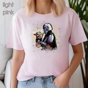 K&ouml;nnte beinhalten: Ein hellrosa T-Shirt mit einem Aquarell-Bild eines Mandalorianer-Charakters, der ein kleines gr&uuml;nes au&szlig;erirdisches Wesen h&auml;lt.