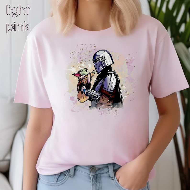 K&ouml;nnte beinhalten: Ein hellrosa T-Shirt mit einem Aquarell-Bild eines Mandalorianer-Charakters, der ein kleines gr&uuml;nes au&szlig;erirdisches Wesen h&auml;lt.
