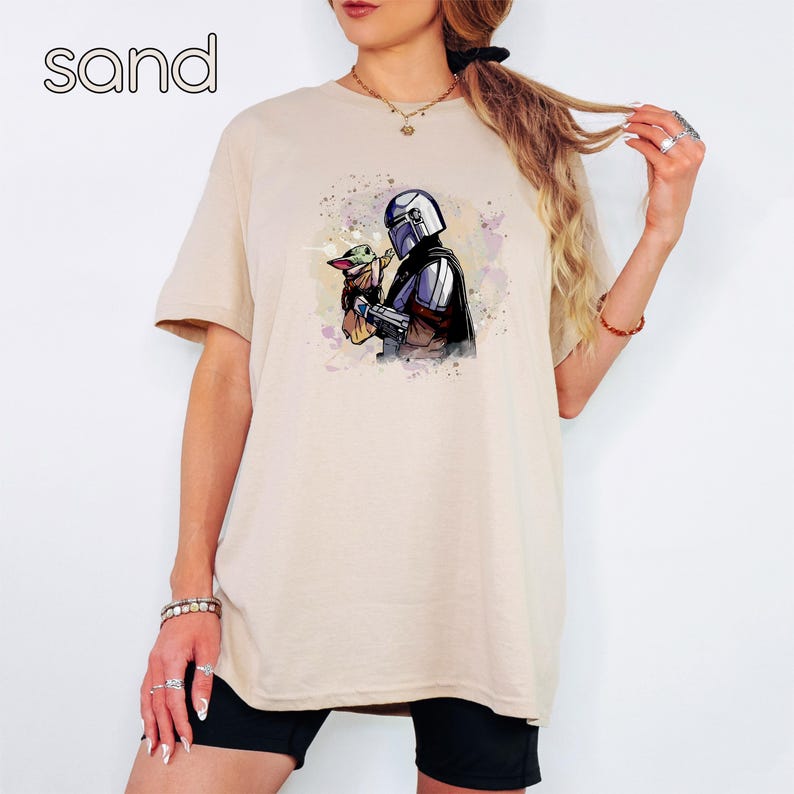K&ouml;nnte beinhalten: Ein beiges T-Shirt mit einer Aquarell-Illustration eines Mandalorianer-Charakters, der ein kleines, gr&uuml;nes Wesen h&auml;lt. Die Illustration ist in Blaut&ouml;nen, Gr&uuml;nt&ouml;nen und Violett.