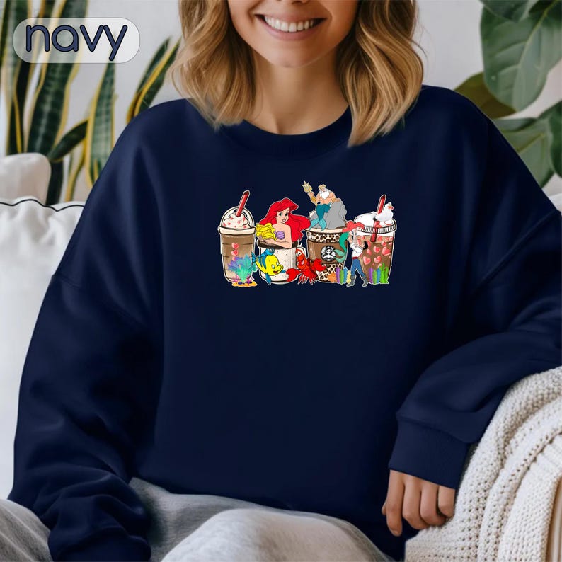 May include: A navy blue sweatshirt with a graphic of Ariel, Ursula, and other characters from The Little Mermaid, all holding coffee cups. The text "Coffee & Mermaids" is printed on the sweatshirt.