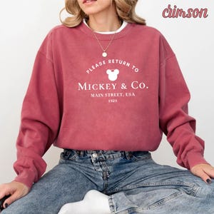 May include: A crimson sweatshirt with a white graphic that says "Please Return To Mickey & Co. Main Street, USA 1928" with a white Mickey Mouse head silhouette.