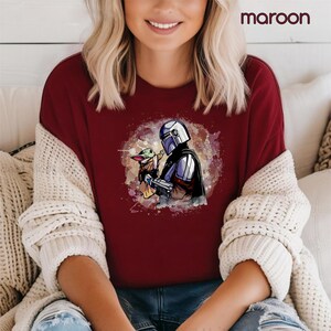 K&ouml;nnte beinhalten: Ein maroonfarbenes T-Shirt mit einem Aquarell-Bild eines Mandalorianer-Charakters, der ein kleines, gr&uuml;nes au&szlig;erirdisches Wesen h&auml;lt. Das Bild ist von einem wei&szlig;en, gesprenkelten Hintergrund umgeben.