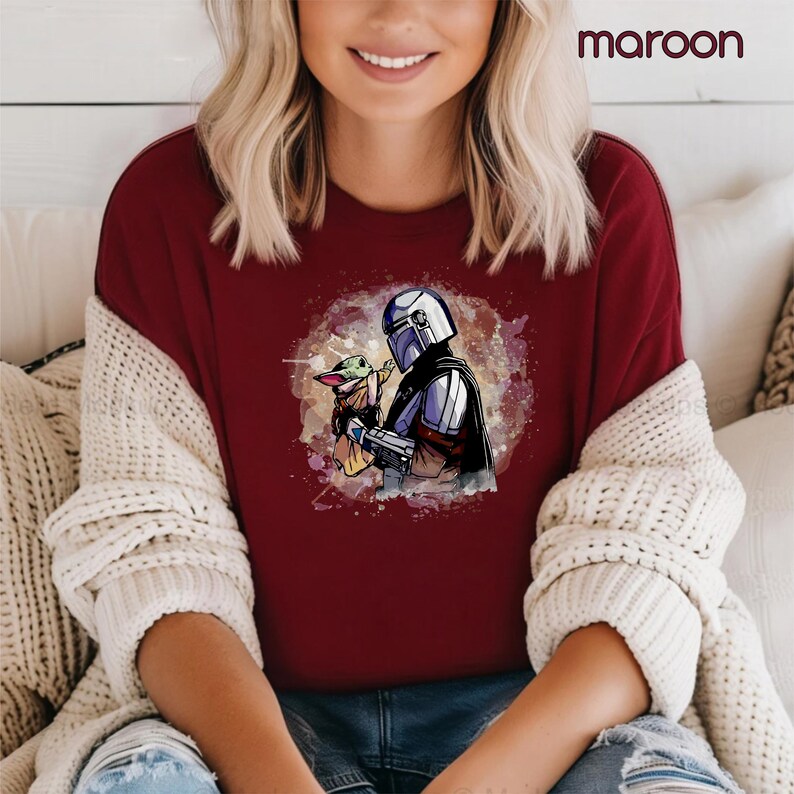 K&ouml;nnte beinhalten: Ein maroonfarbenes T-Shirt mit einem Aquarell-Bild eines Mandalorianer-Charakters, der ein kleines, gr&uuml;nes au&szlig;erirdisches Wesen h&auml;lt. Das Bild ist von einem wei&szlig;en, gesprenkelten Hintergrund umgeben.