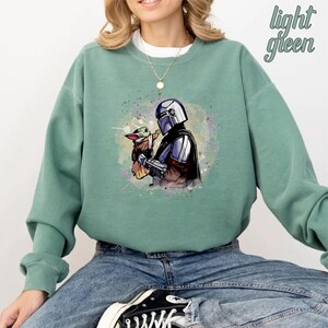 May include: A light green sweatshirt with a watercolor-style image of a Mandalorian character holding a small, green alien creature. The image is centered on the sweatshirt.