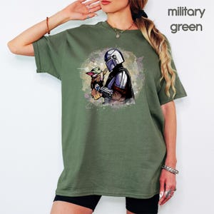 K&ouml;nnte beinhalten: Ein gr&uuml;nes T-Shirt mit einem Aquarell-Bild eines Mandalorianer-Charakters, der ein kleines, gr&uuml;nes au&szlig;erirdisches Wesen h&auml;lt. Der Text "Milit&auml;rgr&uuml;n" ist in der oberen rechten Ecke des Bildes sichtbar.