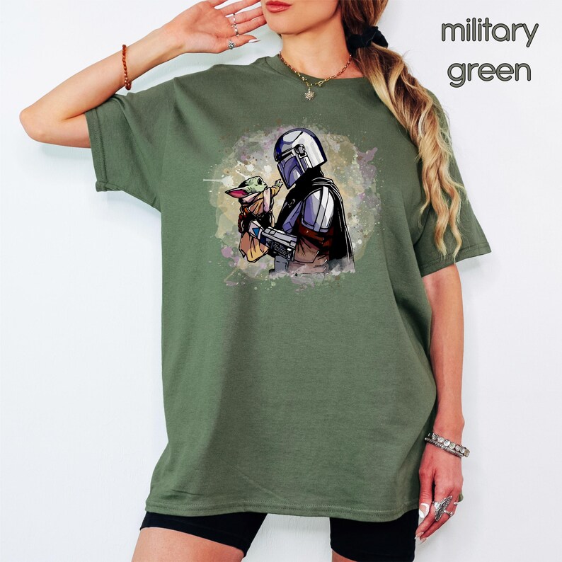 K&ouml;nnte beinhalten: Ein gr&uuml;nes T-Shirt mit einem Aquarell-Bild eines Mandalorianer-Charakters, der ein kleines, gr&uuml;nes au&szlig;erirdisches Wesen h&auml;lt. Der Text "Milit&auml;rgr&uuml;n" ist in der oberen rechten Ecke des Bildes sichtbar.