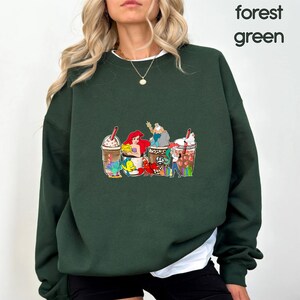 May include: A forest green sweatshirt with a graphic print of characters from the Little Mermaid enjoying a coffee drink. The characters include Ariel, Flounder, Sebastian, and Ursula.