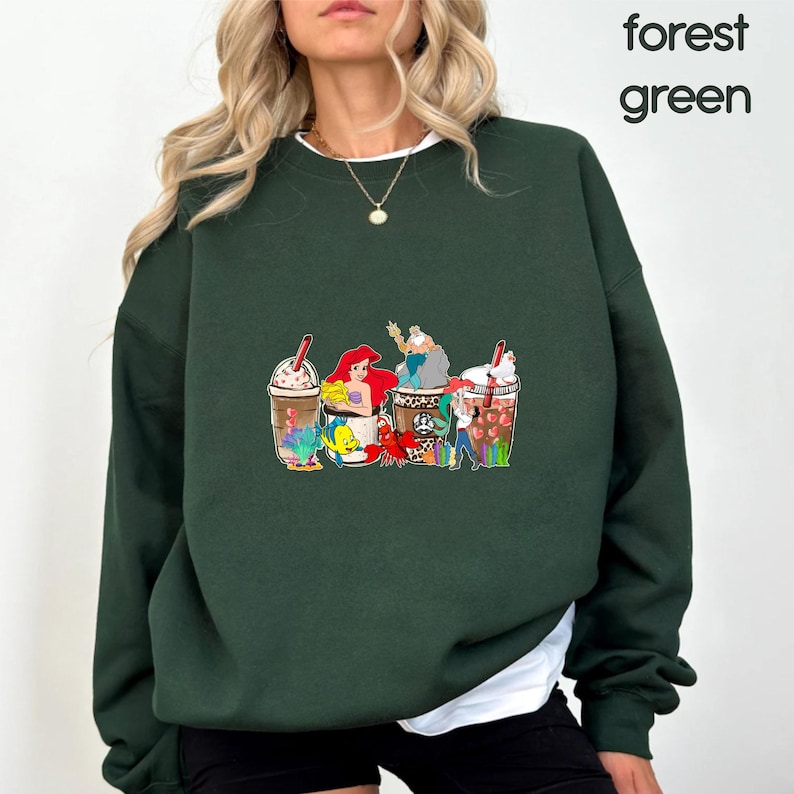 May include: A forest green sweatshirt with a graphic print of characters from the Little Mermaid enjoying a coffee drink. The characters include Ariel, Flounder, Sebastian, and Ursula.