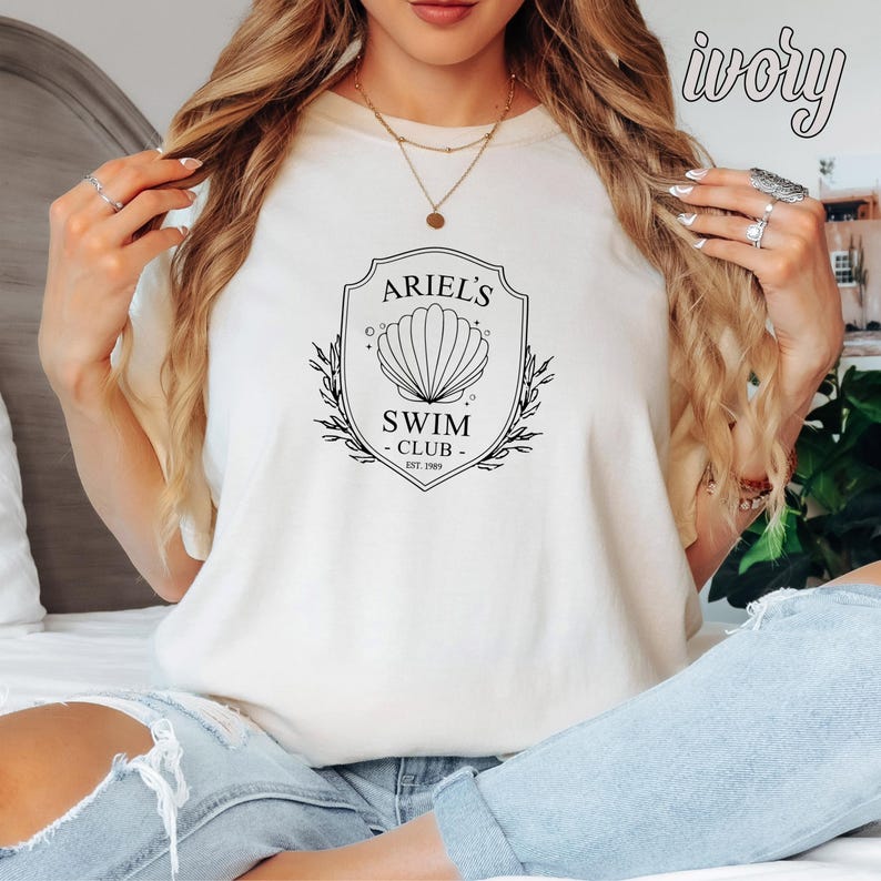 May include: A white t-shirt with a black graphic design featuring a seashell and the text "Ariel's Swim Club Est. 1989".