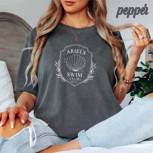May include: A gray t-shirt with a white graphic design that says "Ariel's Swim Club Est. 2003" with a seashell and floral accents.