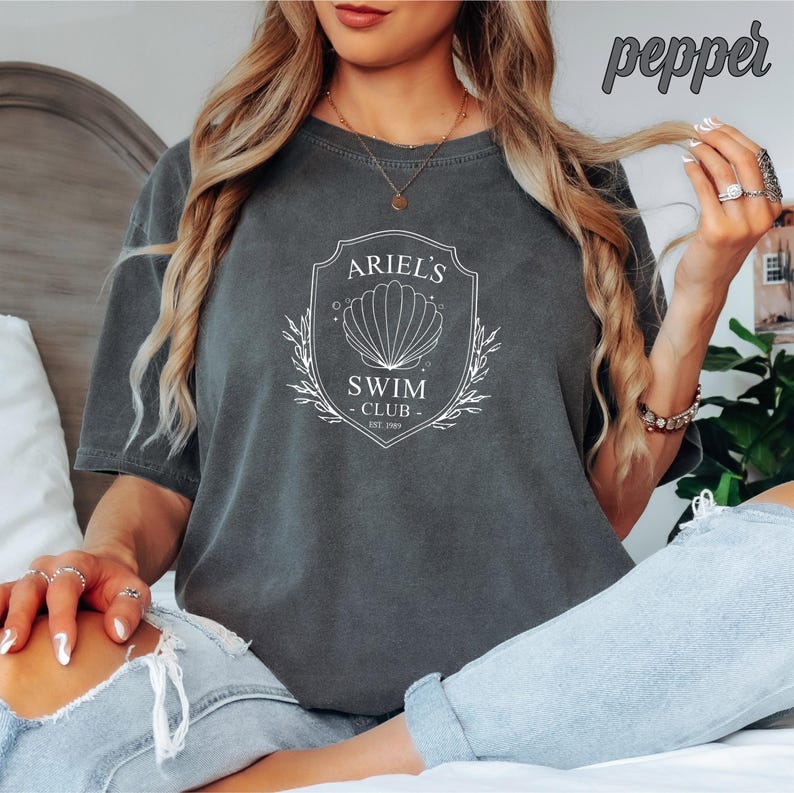 May include: A gray t-shirt with a white graphic design that says "Ariel's Swim Club Est. 2003" with a seashell and floral accents.