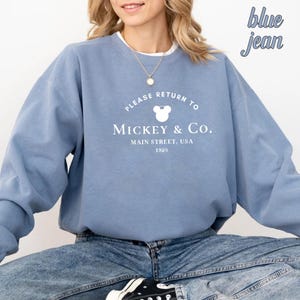 May include: A light blue sweatshirt with a white graphic that says "Please Return to Mickey & Co. Main Street, USA 1928" with a Mickey Mouse head silhouette. The model is wearing blue jeans.