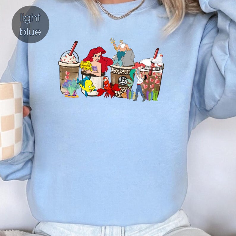 May include: Light blue sweatshirt with a graphic of Disney characters from The Little Mermaid and The Princess and the Frog, including Ariel, Ursula, Prince Eric, Tiana, and Naveen, all depicted as coffee cups.