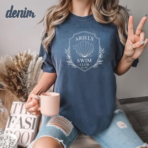 May include: A woman wearing a blue t-shirt with a white graphic design that says "Ariel's Swim Club - Est 1989" and a seashell. She is wearing ripped jeans and holding a pink mug.