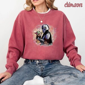 May include: A red sweatshirt with a watercolor-style image of a Mandalorian character holding a small green alien creature. The image is surrounded by a pink and blue watercolor background.