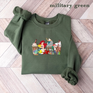 May include: A green sweatshirt with a graphic print of Disney characters, including Ariel, Ursula, and Sebastian, enjoying a cup of coffee. The characters are depicted in a cartoon style and are surrounded by coffee beans and other coffee-related imagery.