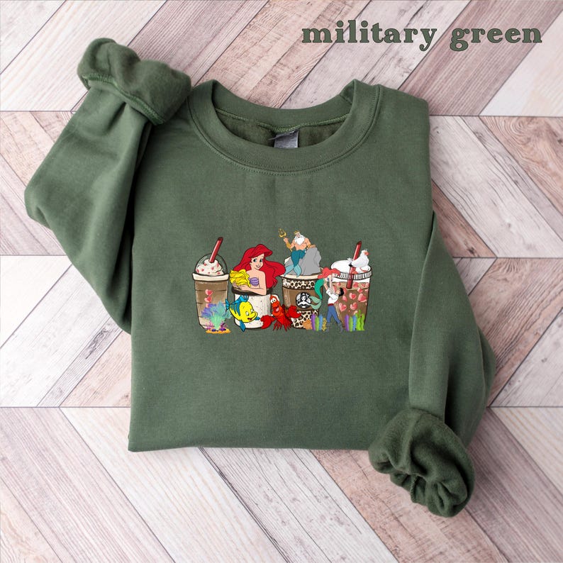 May include: A green sweatshirt with a graphic print of Disney characters, including Ariel, Ursula, and Sebastian, enjoying a cup of coffee. The characters are depicted in a cartoon style and are surrounded by coffee beans and other coffee-related imagery.