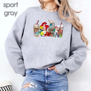 May include: A gray sweatshirt with a graphic of four coffee cups featuring characters from the Disney movie The Little Mermaid. The cups are decorated with Ariel, Ursula, Prince Eric, and Sebastian. The text "sport gray" is in the top left corner of the image.
