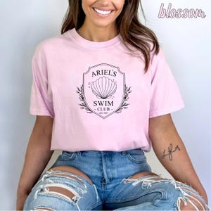 May include: A pink t-shirt with a black graphic design featuring a seashell and the text "Ariel's Swim Club Est. 1989".