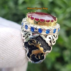 Op de afbeelding: Een zilveren ring met een grote, ovale, rode edelsteen gezet in een gouden bezel. De ring heeft blauwe edelstenen en een gedetailleerd hertenontwerp. De tekst "Diamond Luxury Rings" is zichtbaar.