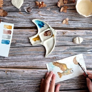 Op de afbeelding: Een persoon schildert een aquarel-illustratie van een bruine en witte uil die vliegt met een halve maan op de achtergrond. De kunstenaar gebruikt een palet met blauwe, bruine en gele verf.