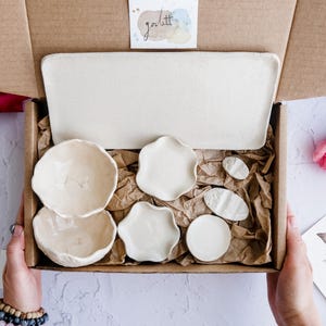 May include: A craft box containing a set of handmade, cream-coloured ceramic dishes. The set includes a rectangular tray, two small bowls, and several decorative plates in various shapes. The box is lined with brown paper and includes a small watercolour painting and a sticker.